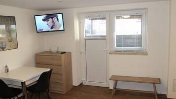 A room with a white table, two black chairs, a wooden bench, a TV on the wall, a door with a window, and a cabinet.