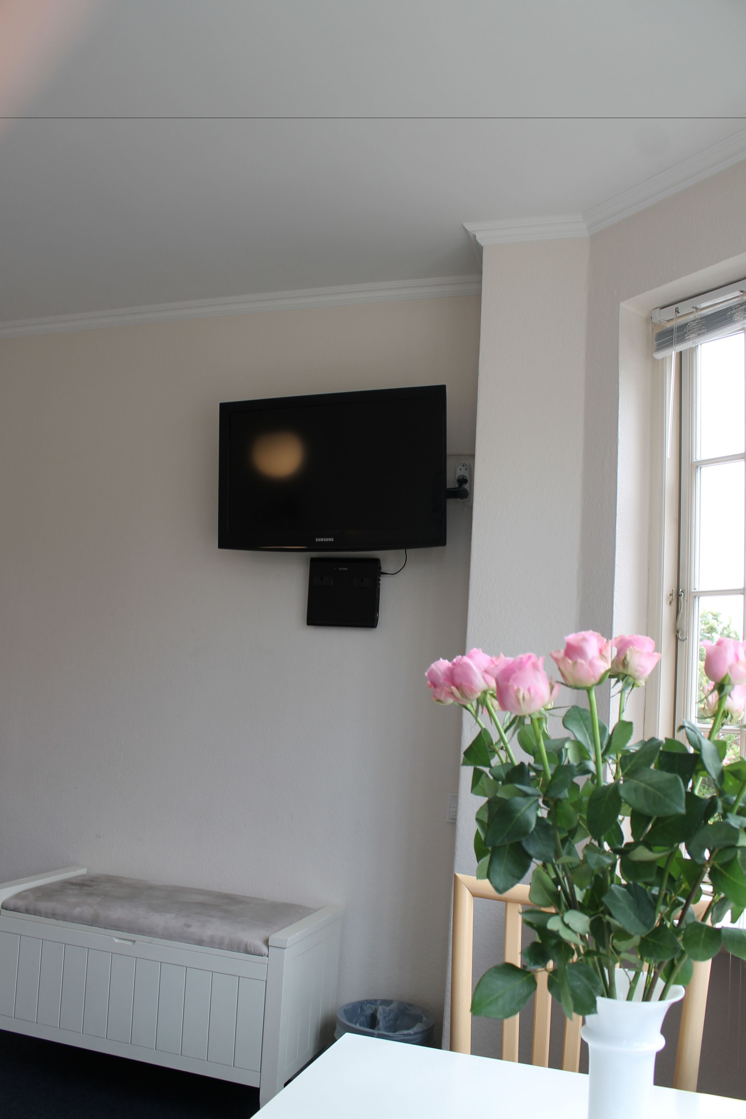 Ein Zimmer mit einer weißen Bank, einem weißen Tisch mit Blumen in einer Vase, einem an der Wand befestigten Fernseher und einem Fenster mit Blick auf Grünflächen.