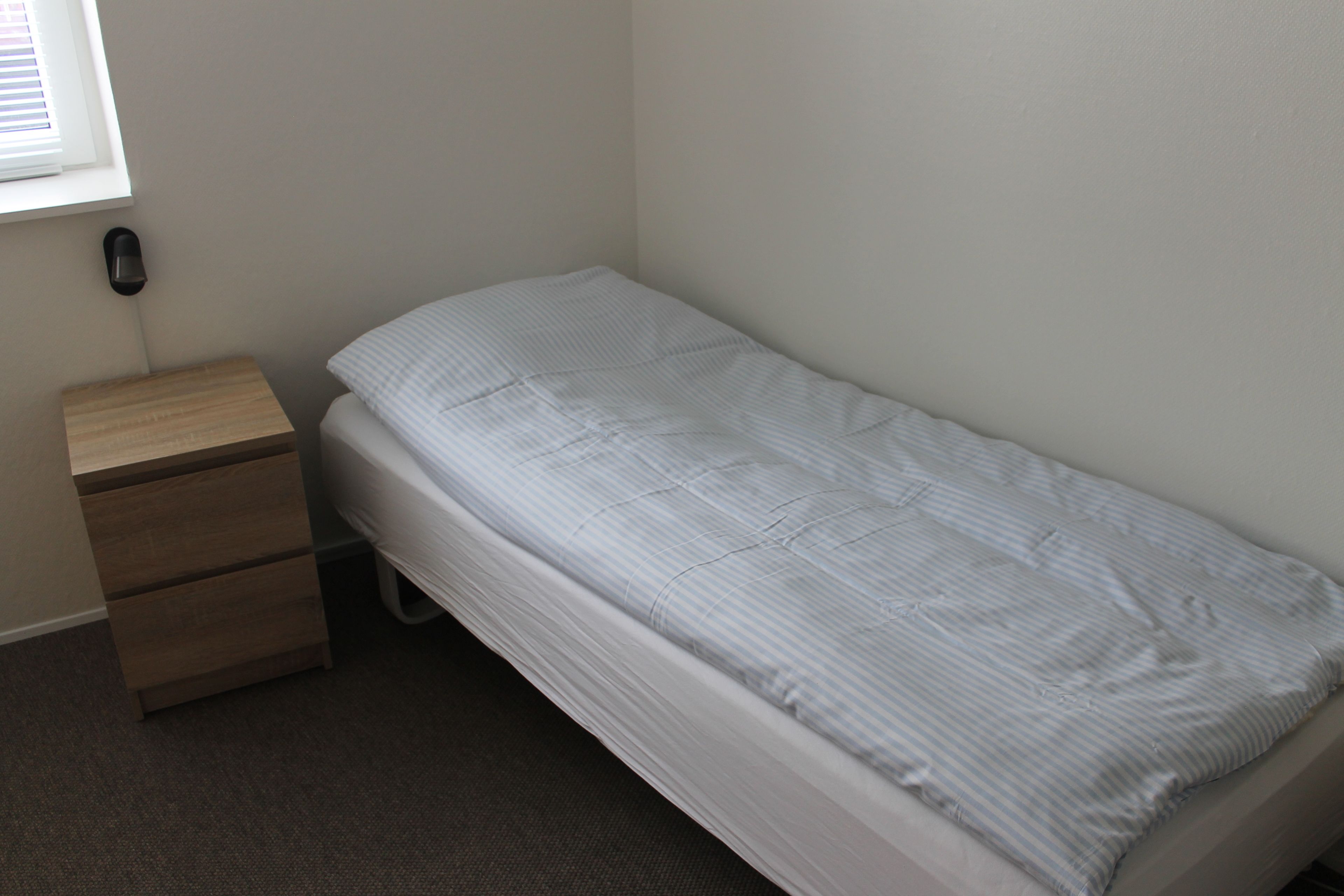 Une petite chambre avec un lit simple, une table de chevet en bois et un téléphone fixé au mur.