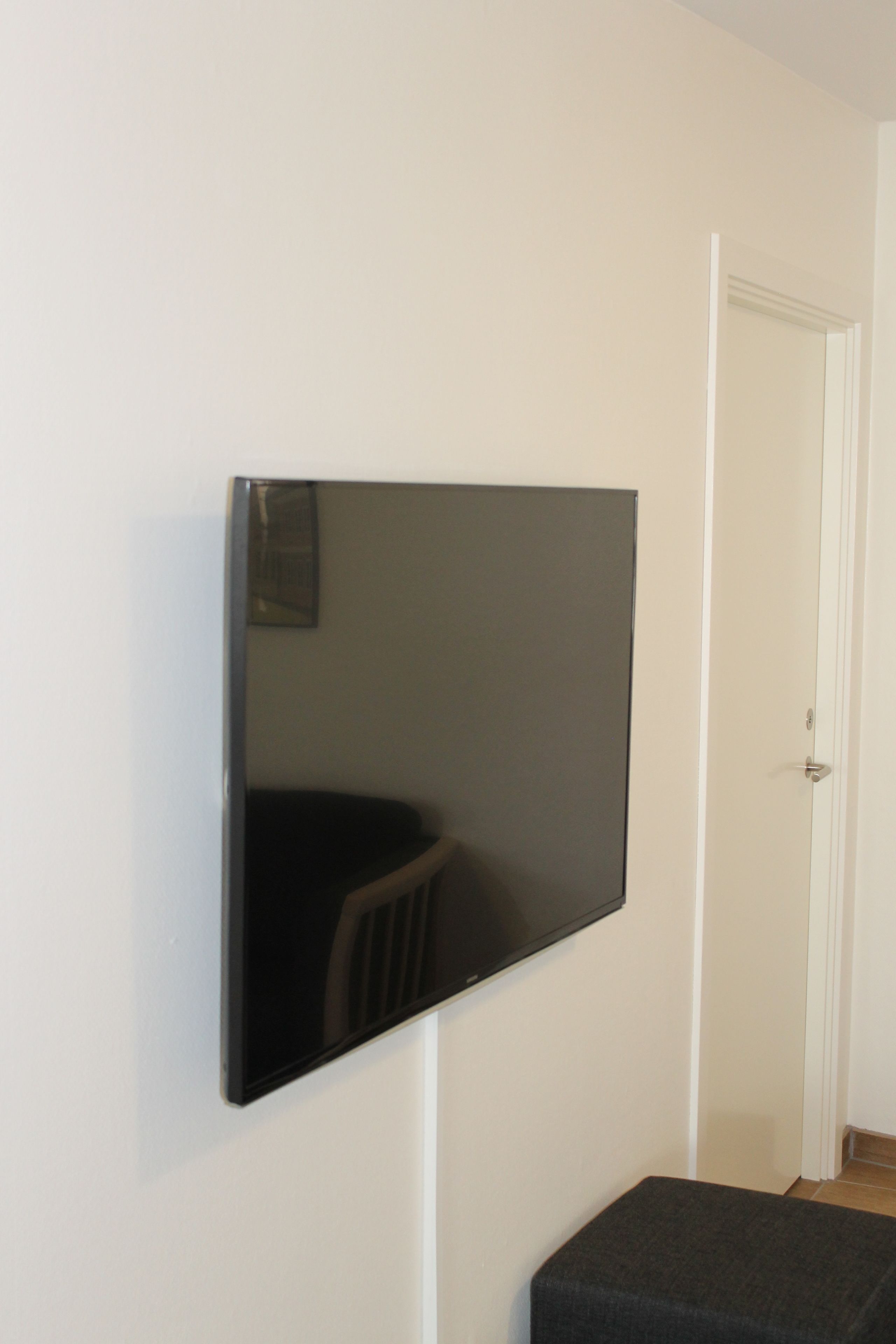 Ein Flachbildfernseher an einer weißen Wand, eine Tür rechts und ein kleiner, dunkel gefärbter Hocker im Vordergrund.