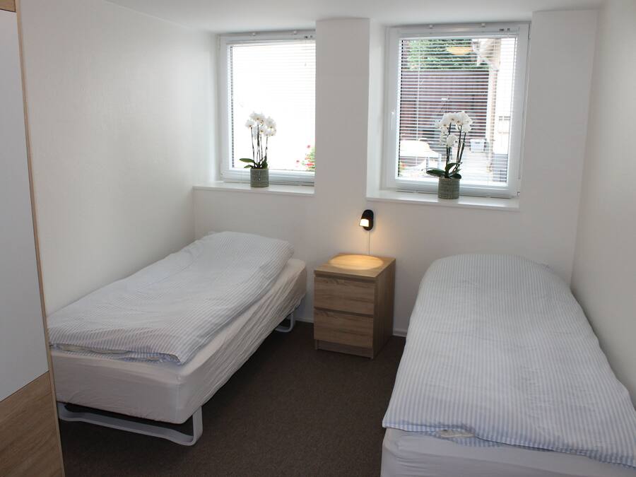 A small, clean room with two single beds, a bedside table, and a window with a view.