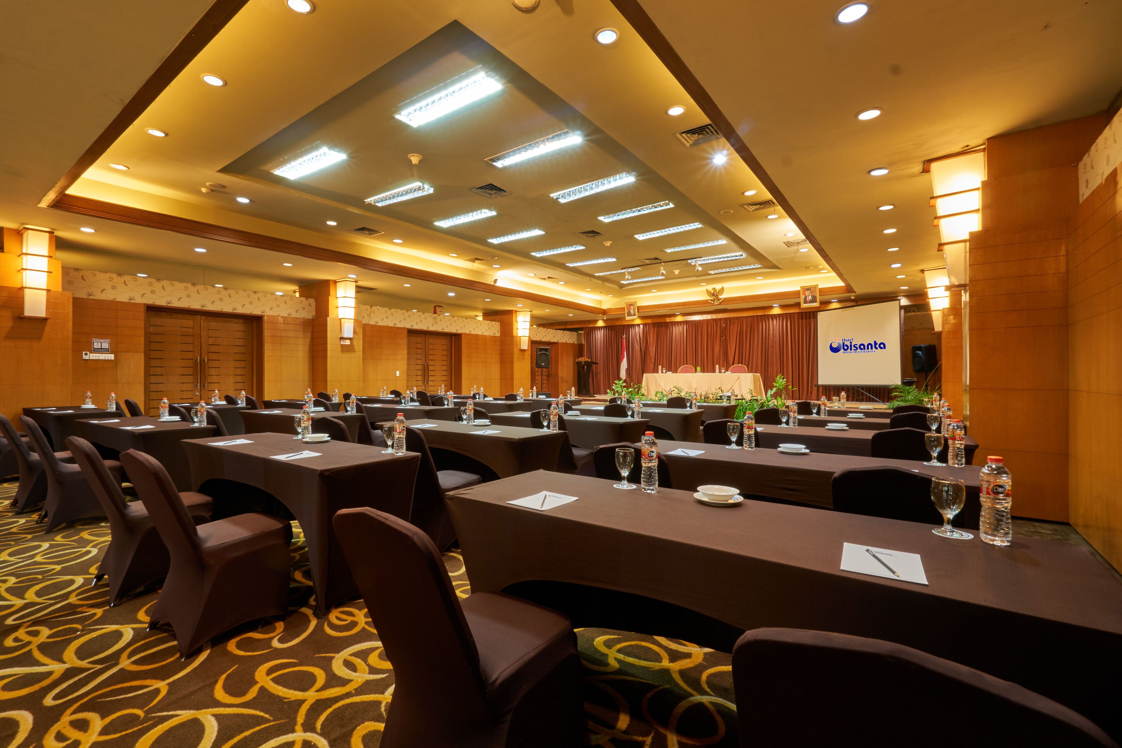A conference room with multiple tables, chairs, and a stage with a screen.
