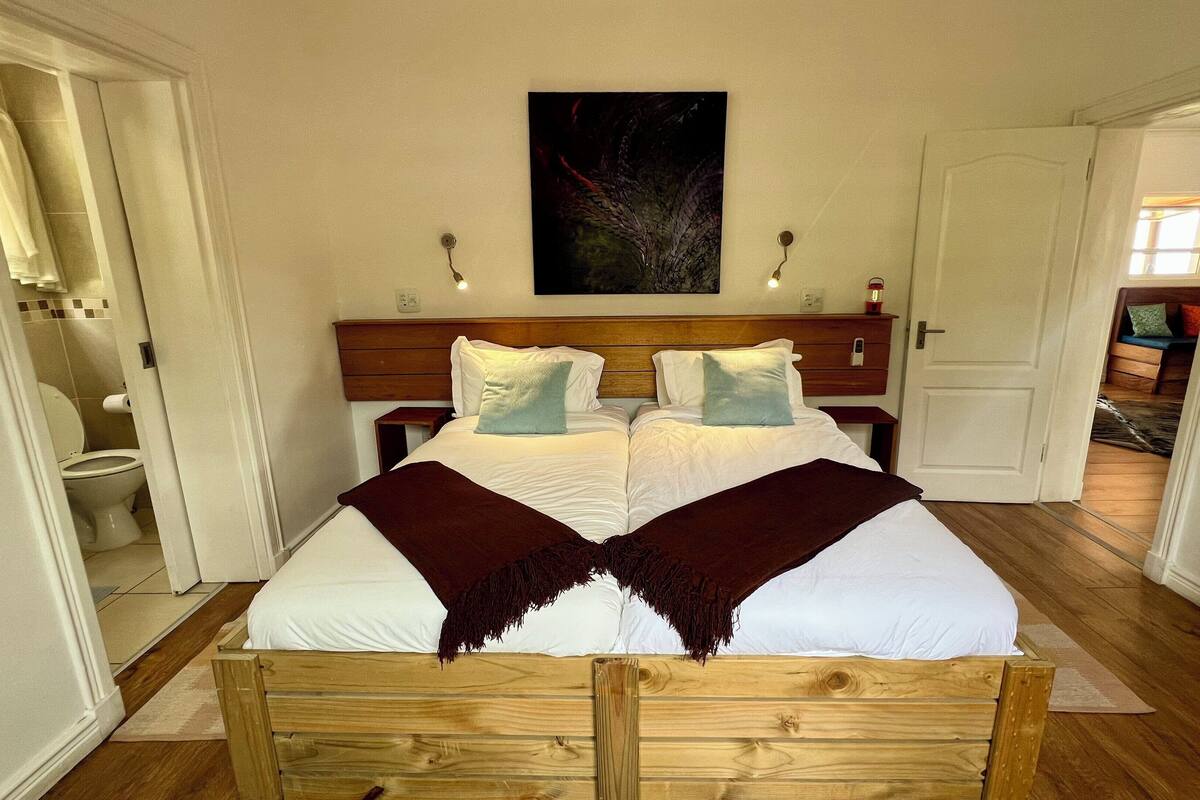 Ein Doppelbett mit Holzkopfstück, zwei Kissen und einer dunklen Tagesdecke, darüber ein Gemälde.