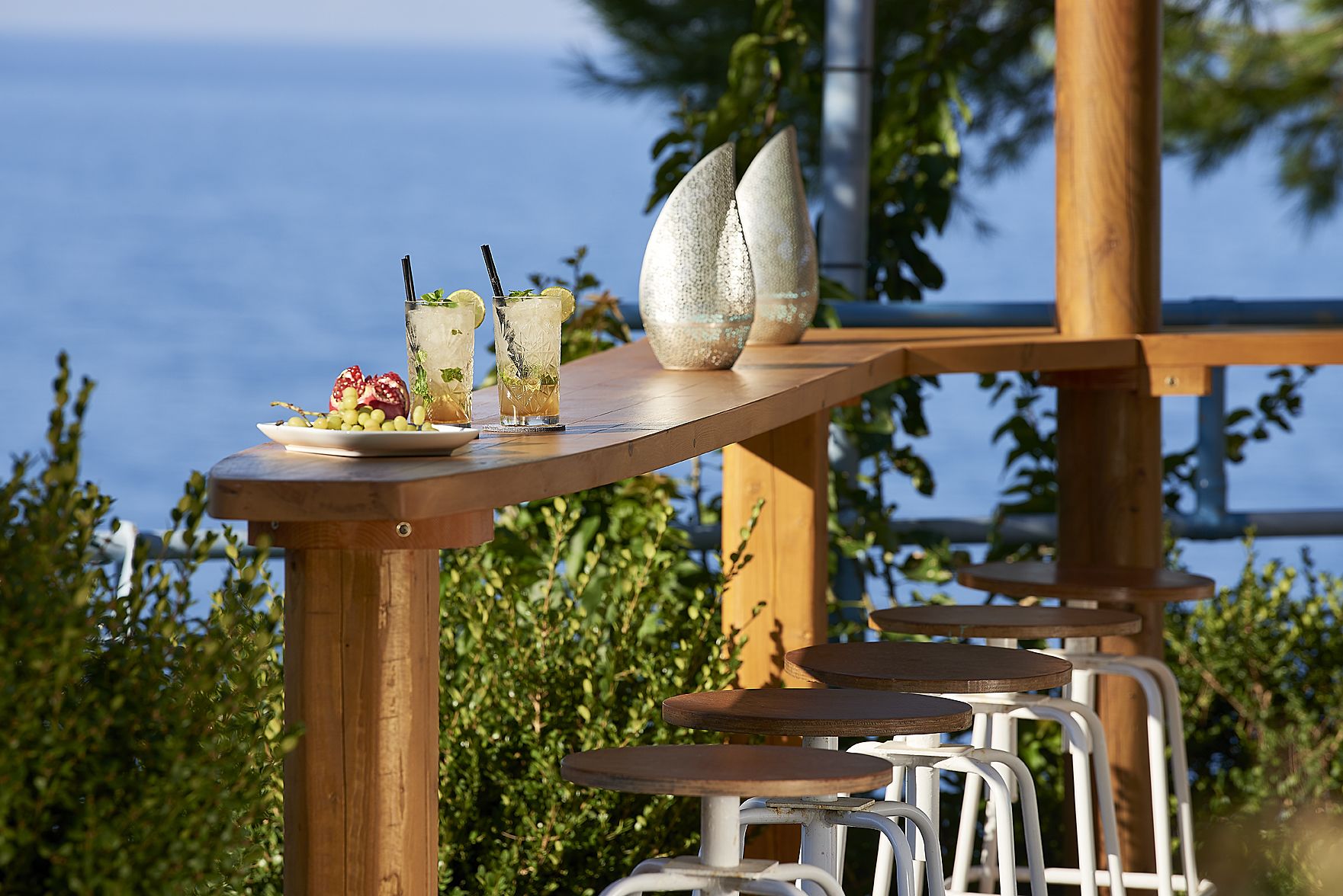Un tavolo di legno con bevande e un piatto di cibo, sullo sfondo del verde e vista sul mare.