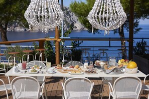 Un área de comedor al aire libre con sillas y mesas de metal blanco, una variedad de comida y bebidas, y luces colgantes blancas decorativas.