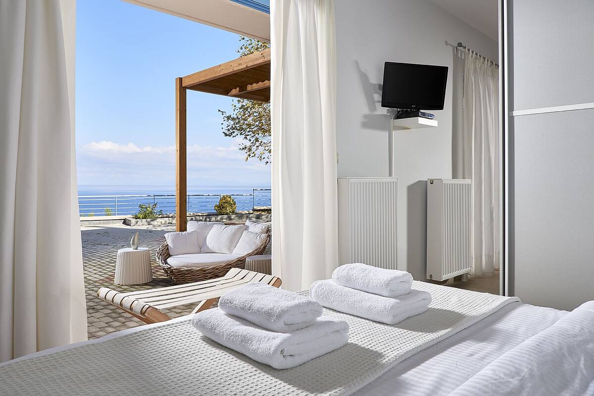 A hotel room with a bed, a TV, a view of the sea, a wicker chair, and a small table.