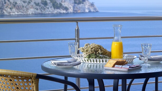 Una mesa con alimentos para el desayuno, un vaso de jugo de naranja y vista al mar.