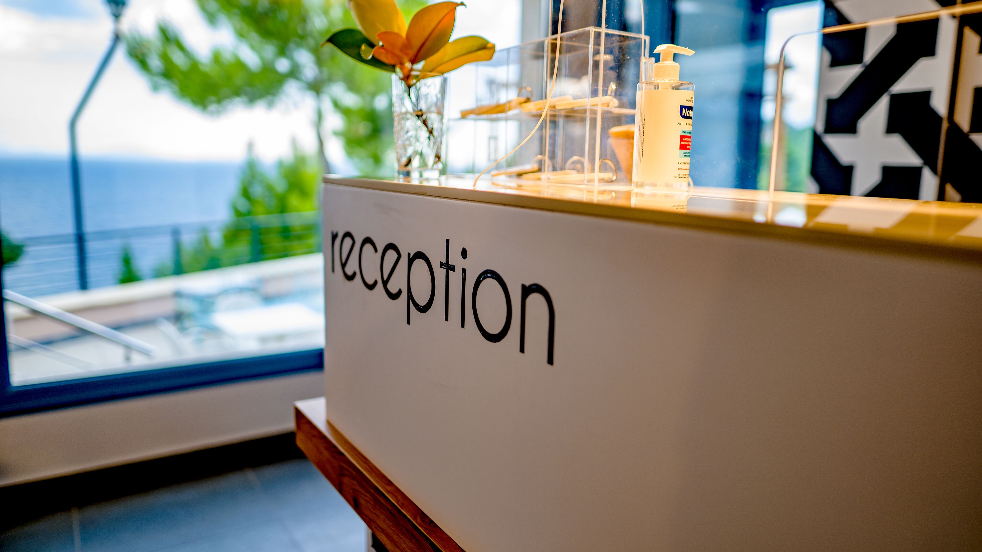 Una mesa de recepción con un letrero que dice 'recepción', un florero con una flor amarilla y un dispensador de gel antibacterial.