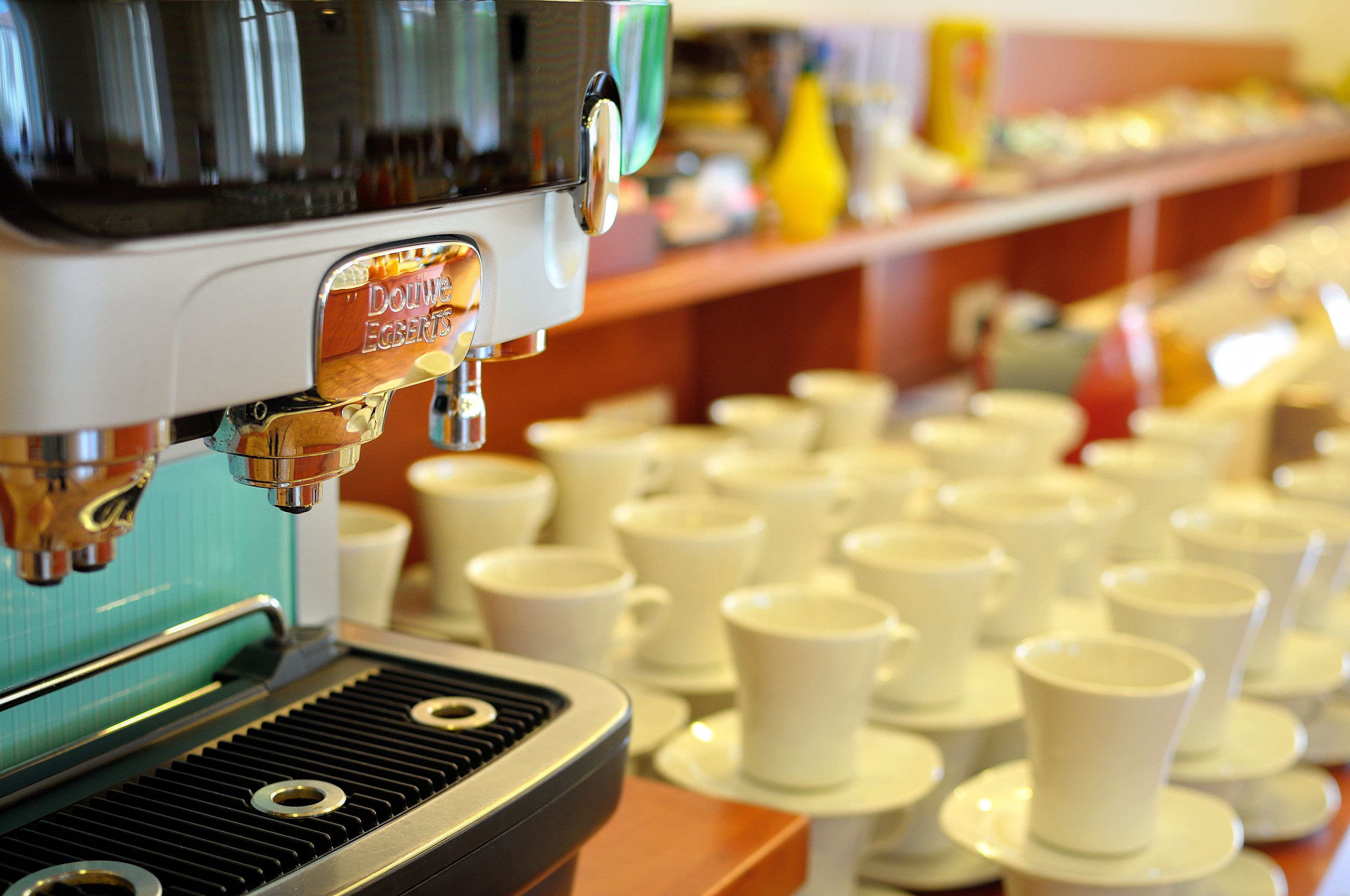 Una cafetera con un rótulo que dice "Douwe Egberts" y una fila de tazas blancas en una bandeja.