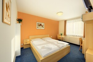 Una habitación de hotel con cama doble, un escritorio, una silla, una mesita, un radiador y una ventana con cortinas.