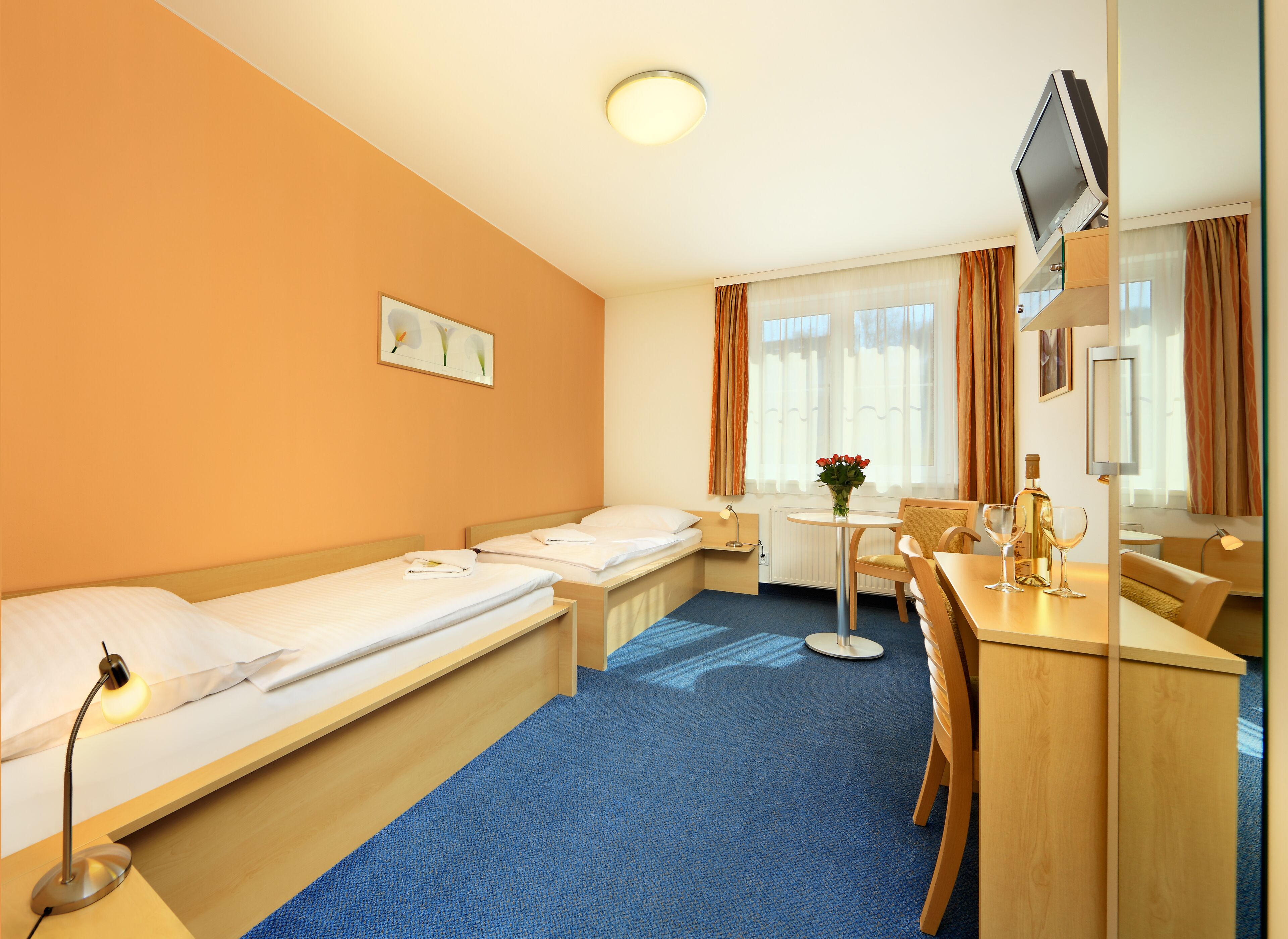 Quarto de hotel com duas camas de solteiro, uma mesinha com um vaso de flores, uma escrivaninha com cadeira, uma televisão e uma janela com cortinas.