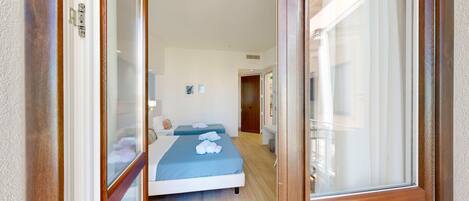 A hotel room with a wooden door, a bed with blue bedding, a bedside table, and a small bedside lamp.