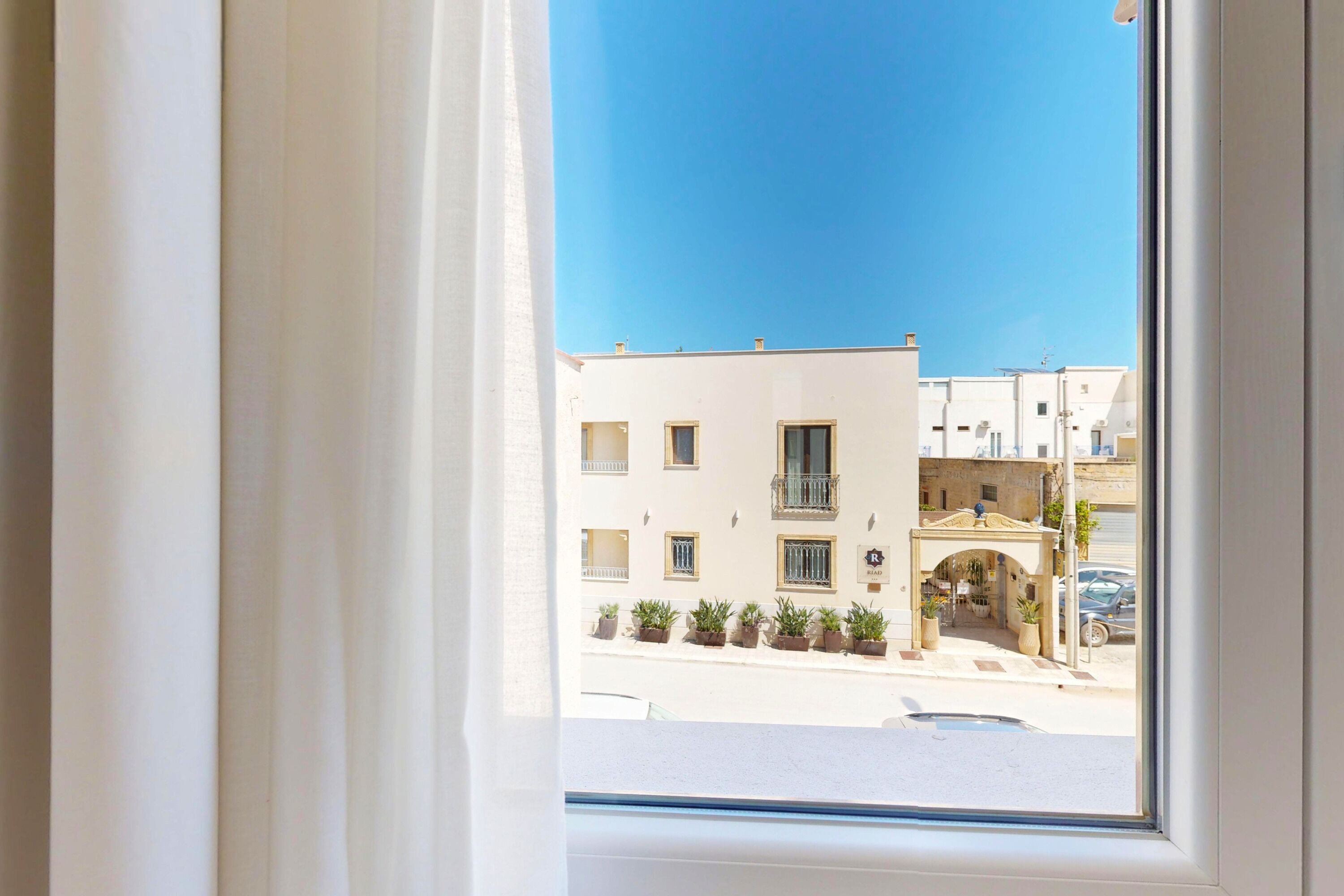 A window view of a building with a balcony, potted plants, and a clear blue sky.