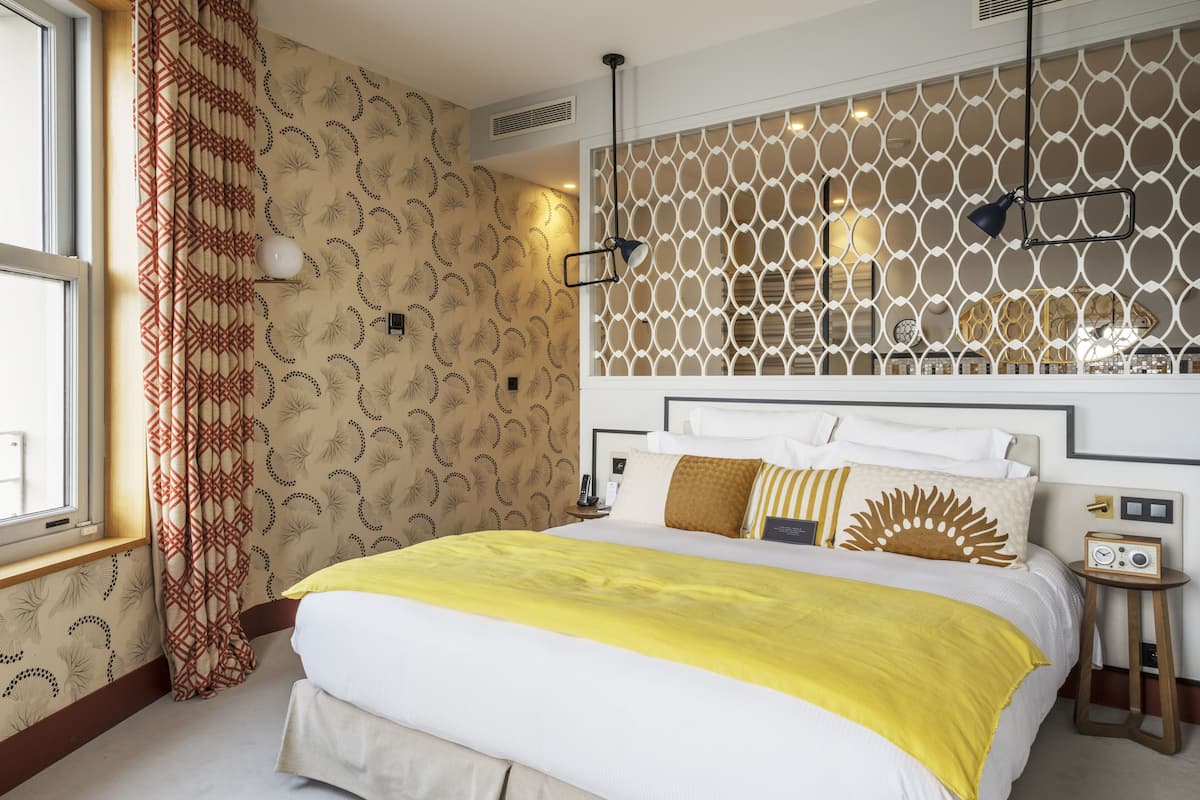 Una habitación de hotel con una cama grande, un diseño decorativo en la pared, una ventana con cortinas y una mesita de noche con una radio.