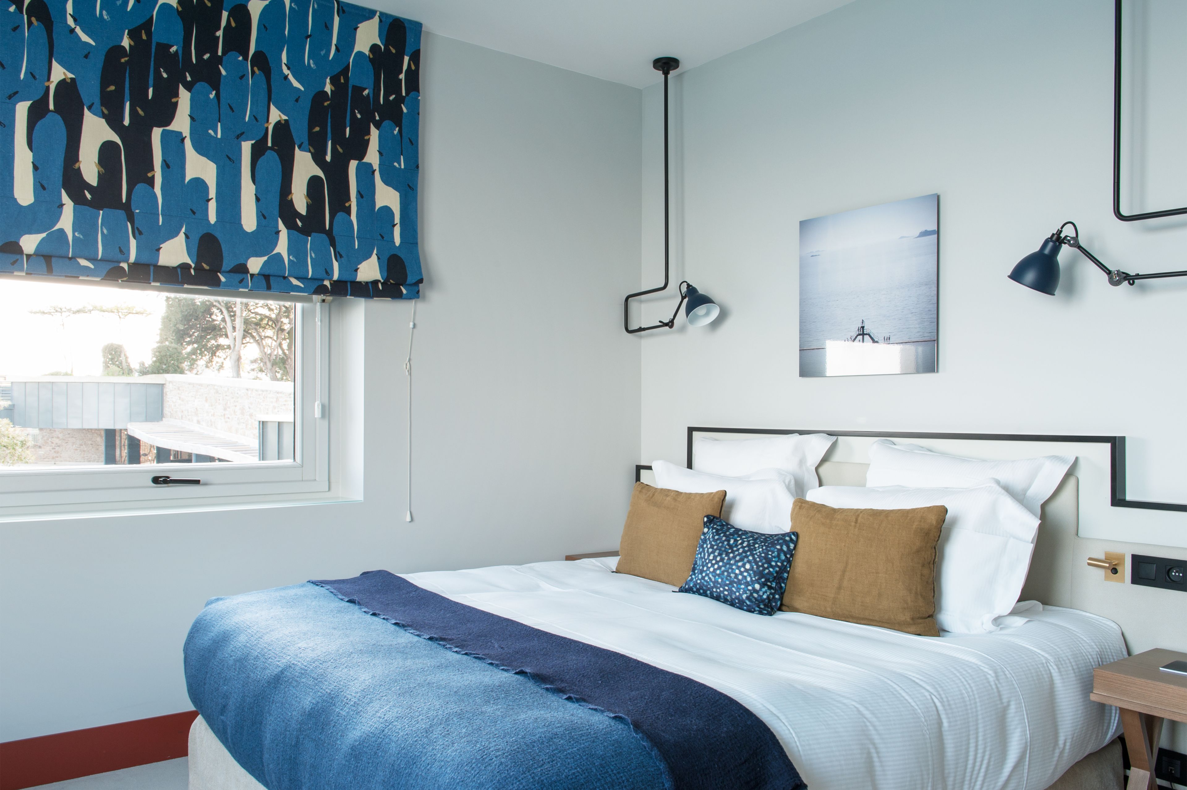 Un dormitorio con cama, ventana, lámpara de pared y un cuadro.