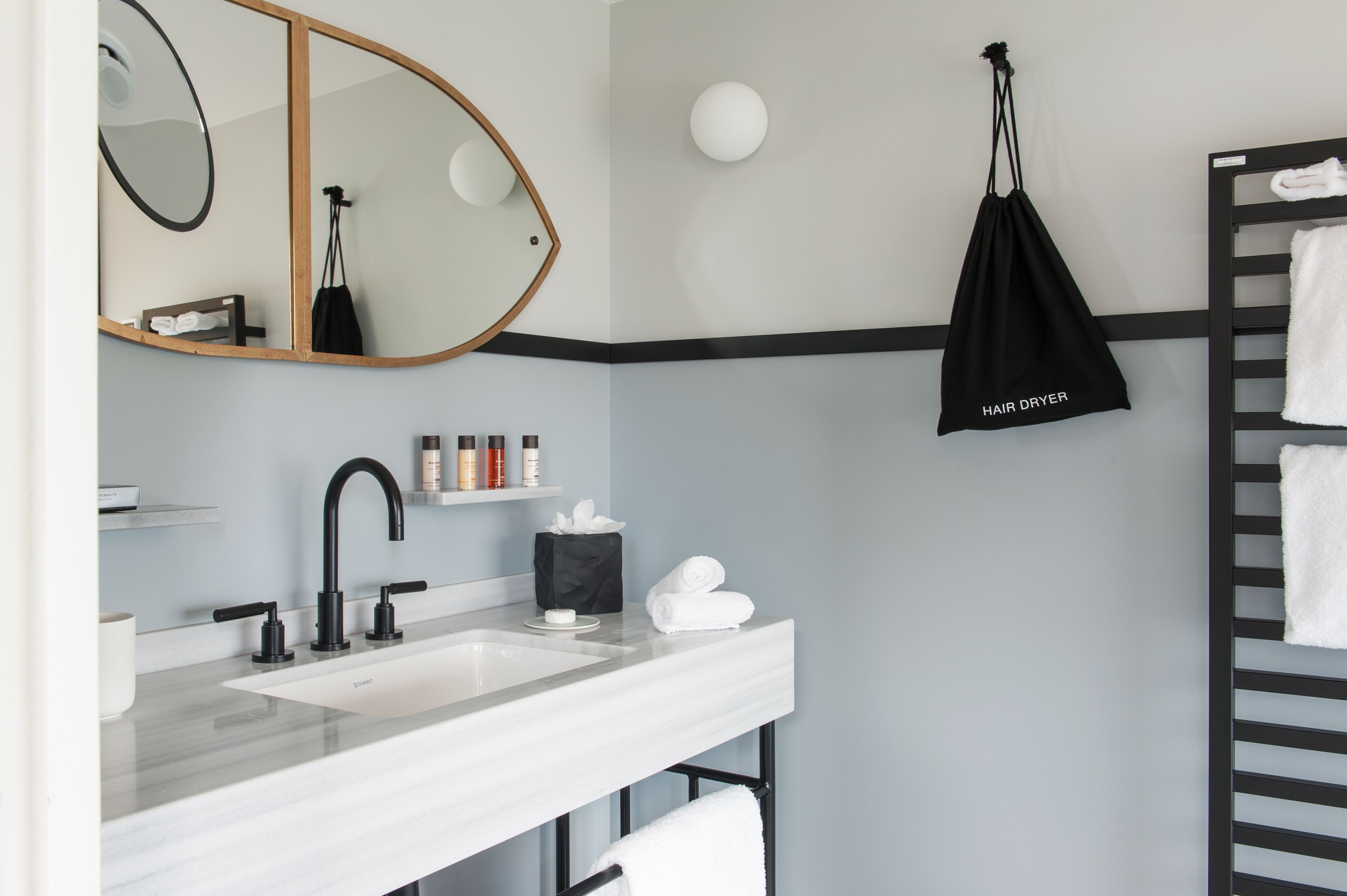 A modern bathroom with a white sink, black faucet, and a mirror with a wooden frame. There is a black bag labeled "HAIR DRYER" hanging on the wall, and a towel rack with white towels.
