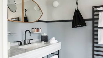 A modern bathroom with a white sink, black faucet, and a mirror with a wooden frame. There is a black bag labeled "HAIR DRYER" hanging on the wall, and a towel rack with white towels.