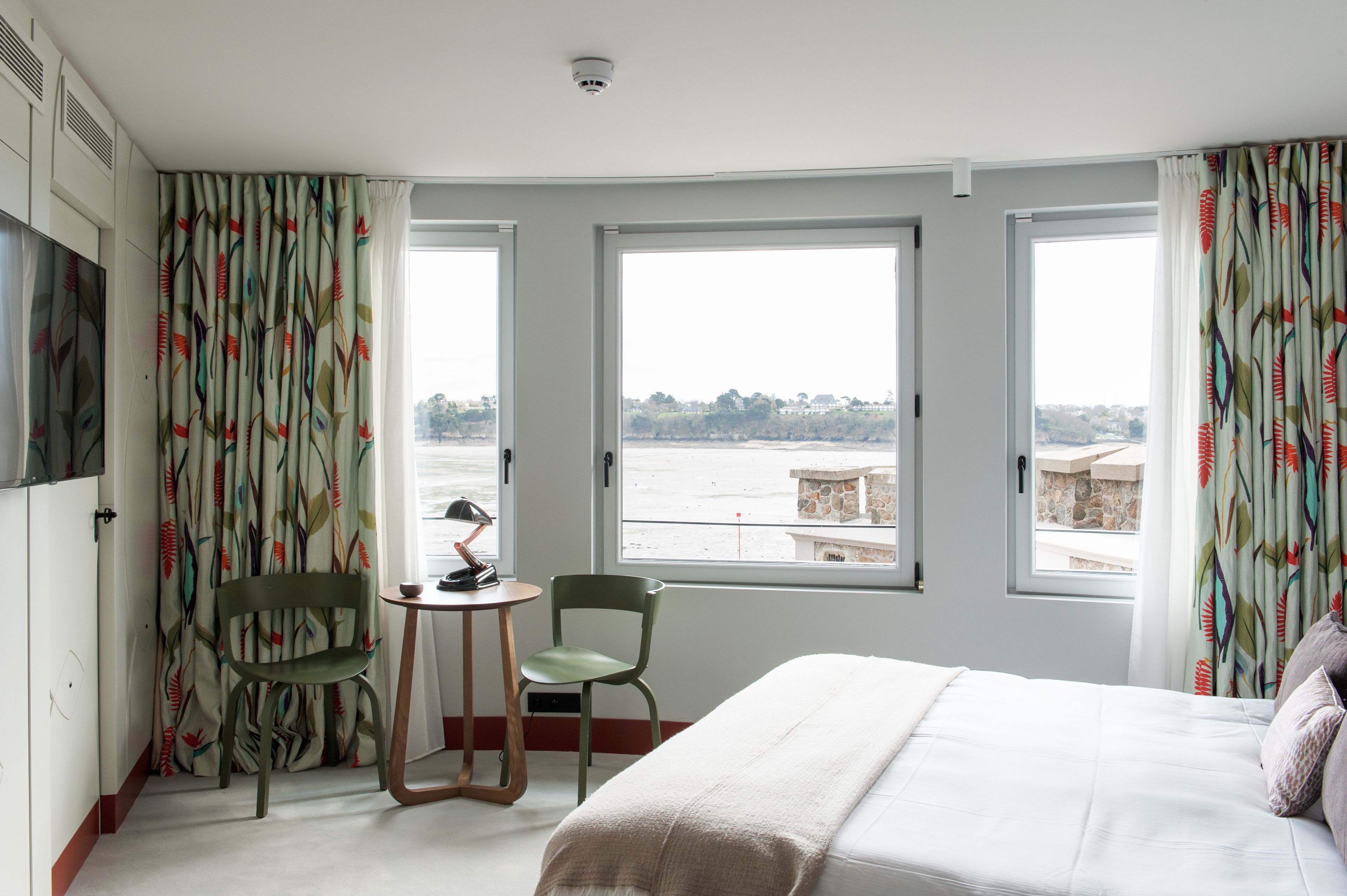 Een hotelkamer met een bed, twee stoelen, een kleine tafel en grote ramen met uitzicht op zee.