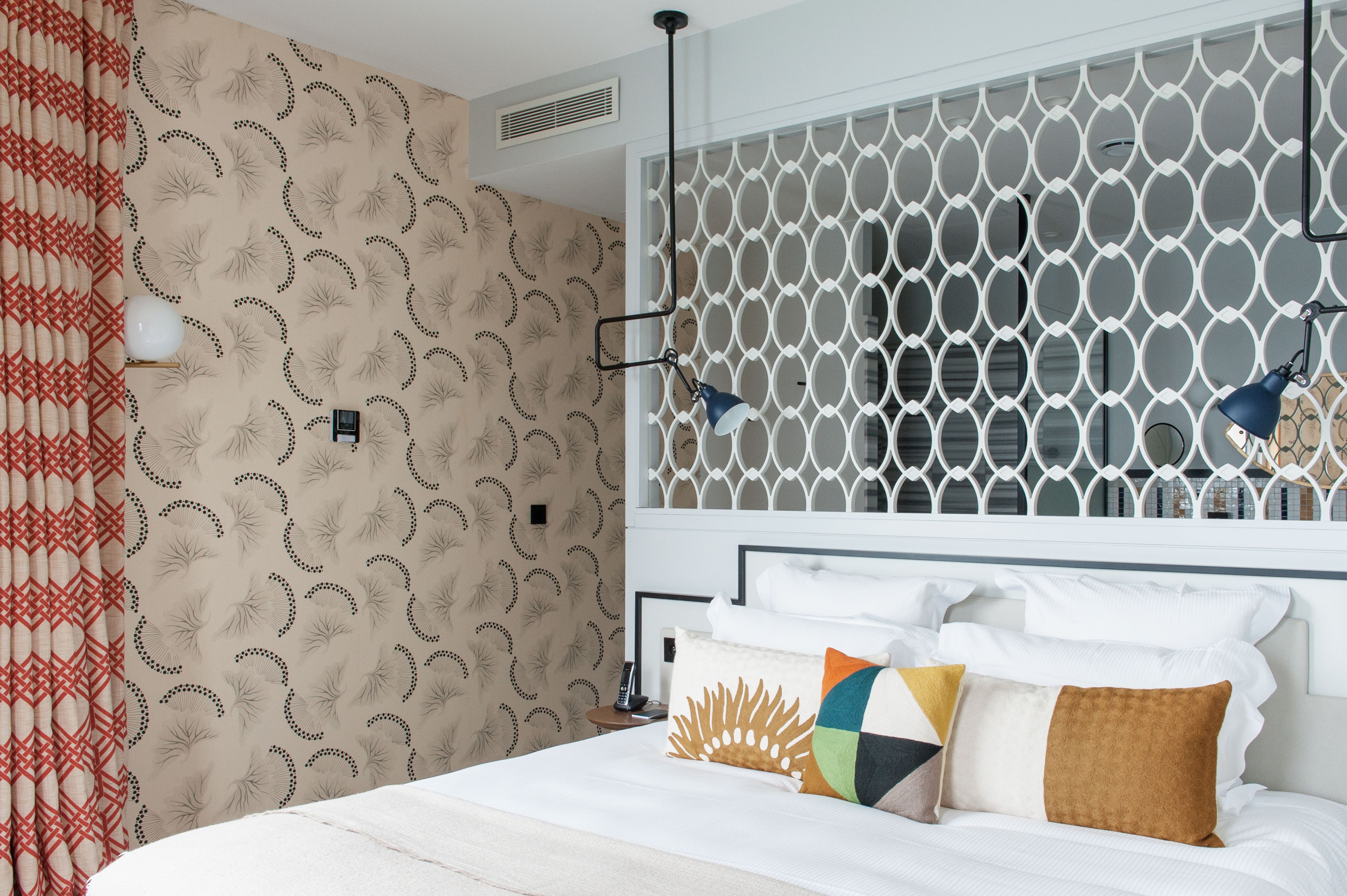 A neatly made bed with white linens and multiple pillows, a patterned wall, and a white headboard with a geometric design.