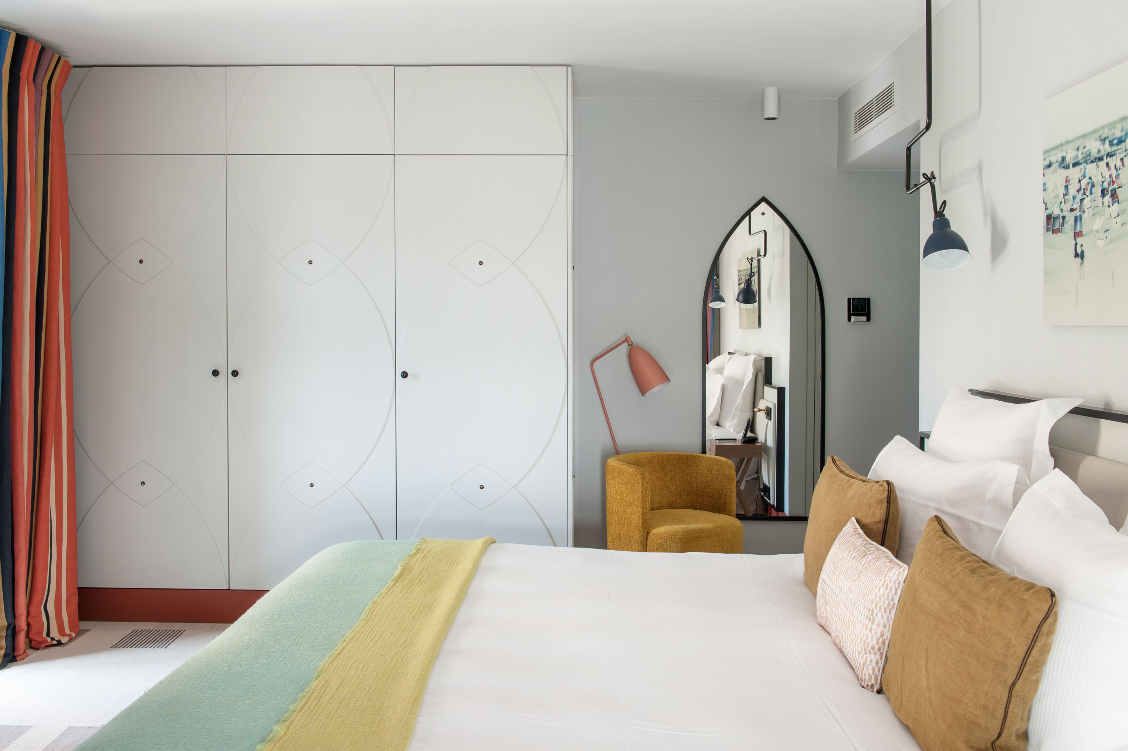 A hotel room with a large bed, a wardrobe, a mirror, and a desk with a lamp.
