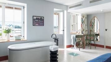 A modern bathroom with a bathtub, a window with a view, a painting, and a mirror with a colorful pattern.