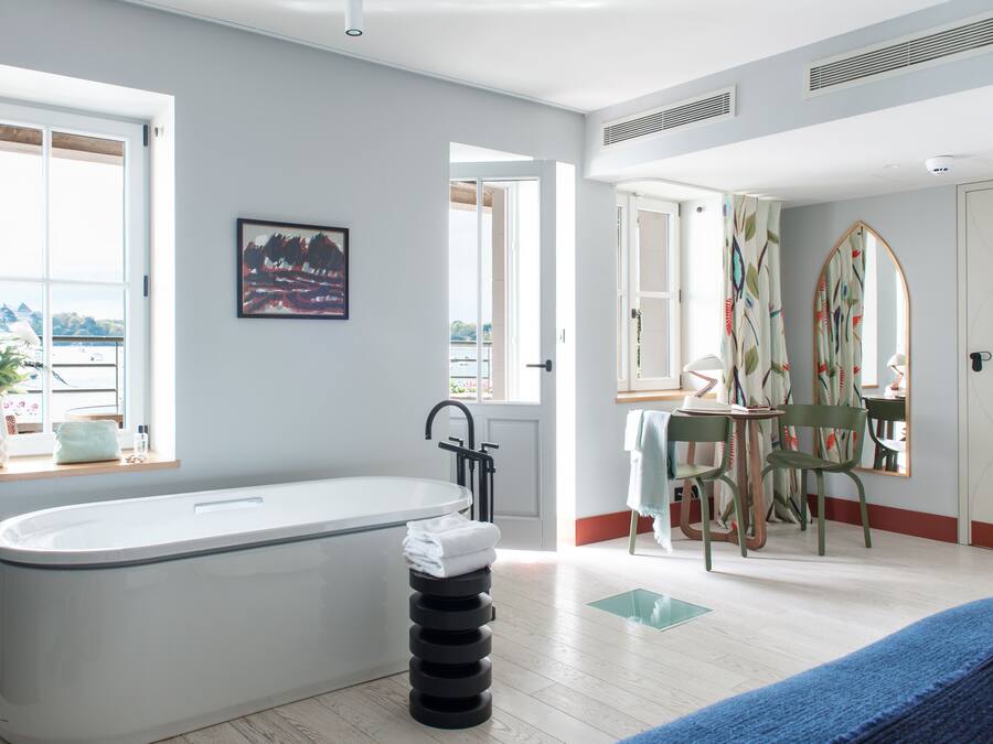 A modern bathroom with a bathtub, a window with a view, a painting, and a mirror with a colorful pattern.