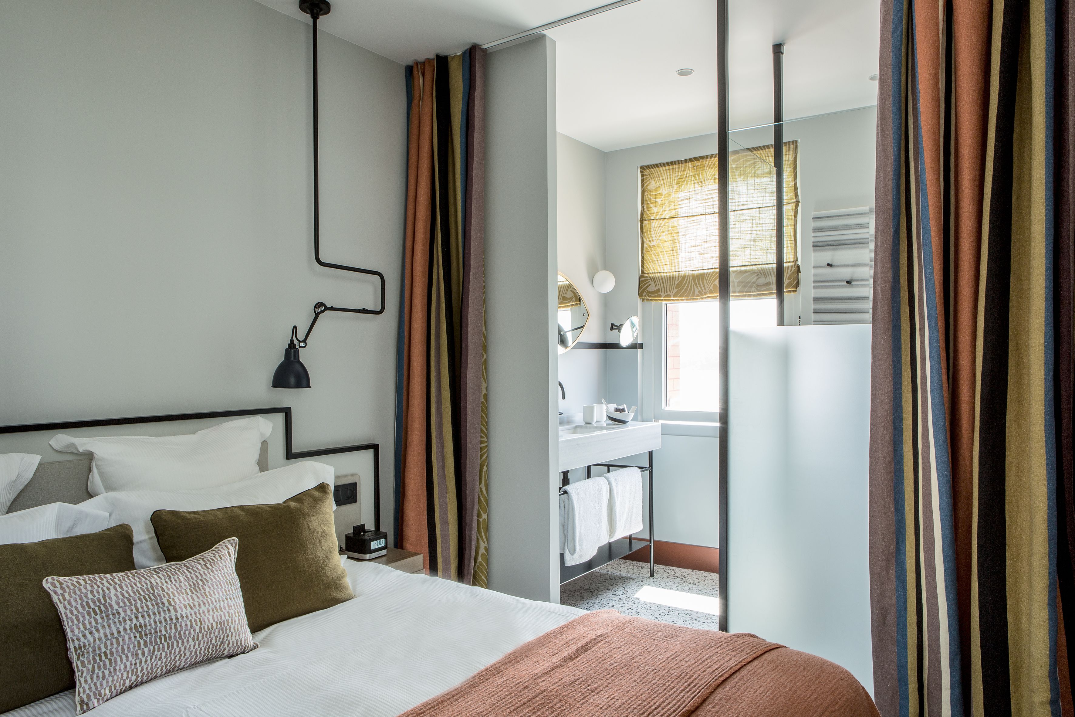 Una habitación de hotel moderna con una cama, una mesita de noche, una lámpara y un baño visible a través de una puerta abierta.