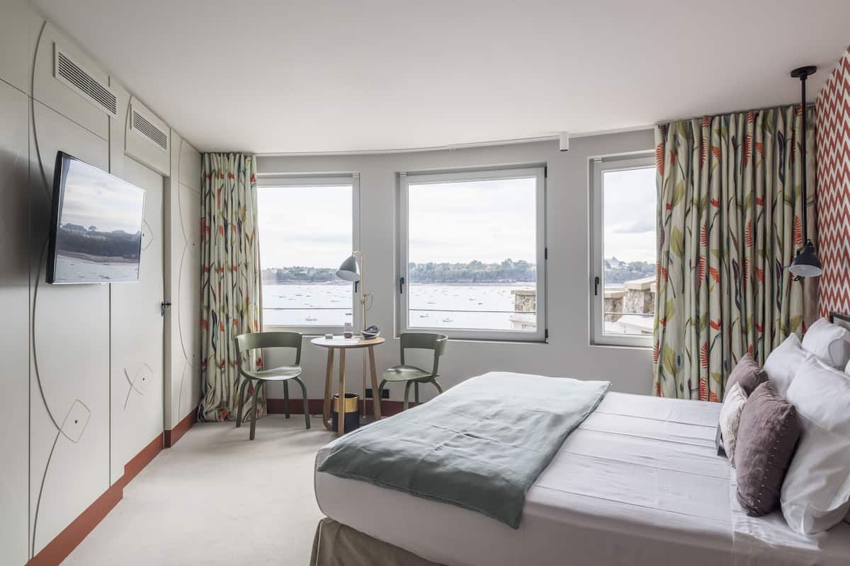 Un cuarto de hotel con cama, televisor montado en la pared, una pequeña mesa con sillas y ventanas amplias con vista al mar.