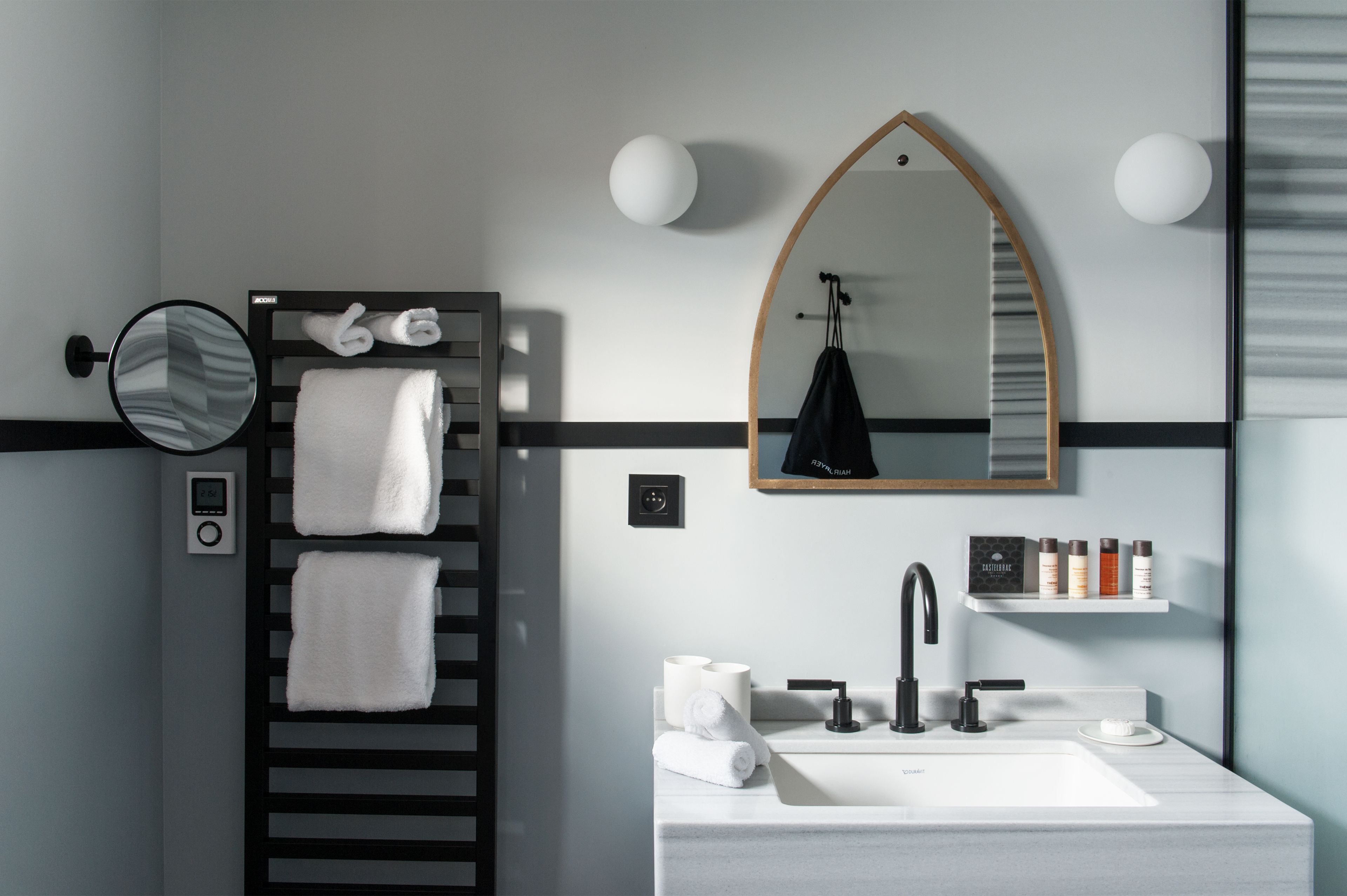 Een moderne badkamer met een zwarte handdoekhouder, een ronde spiegel, een wastafel met een zwarte kraan en een plank met toiletartikelen.