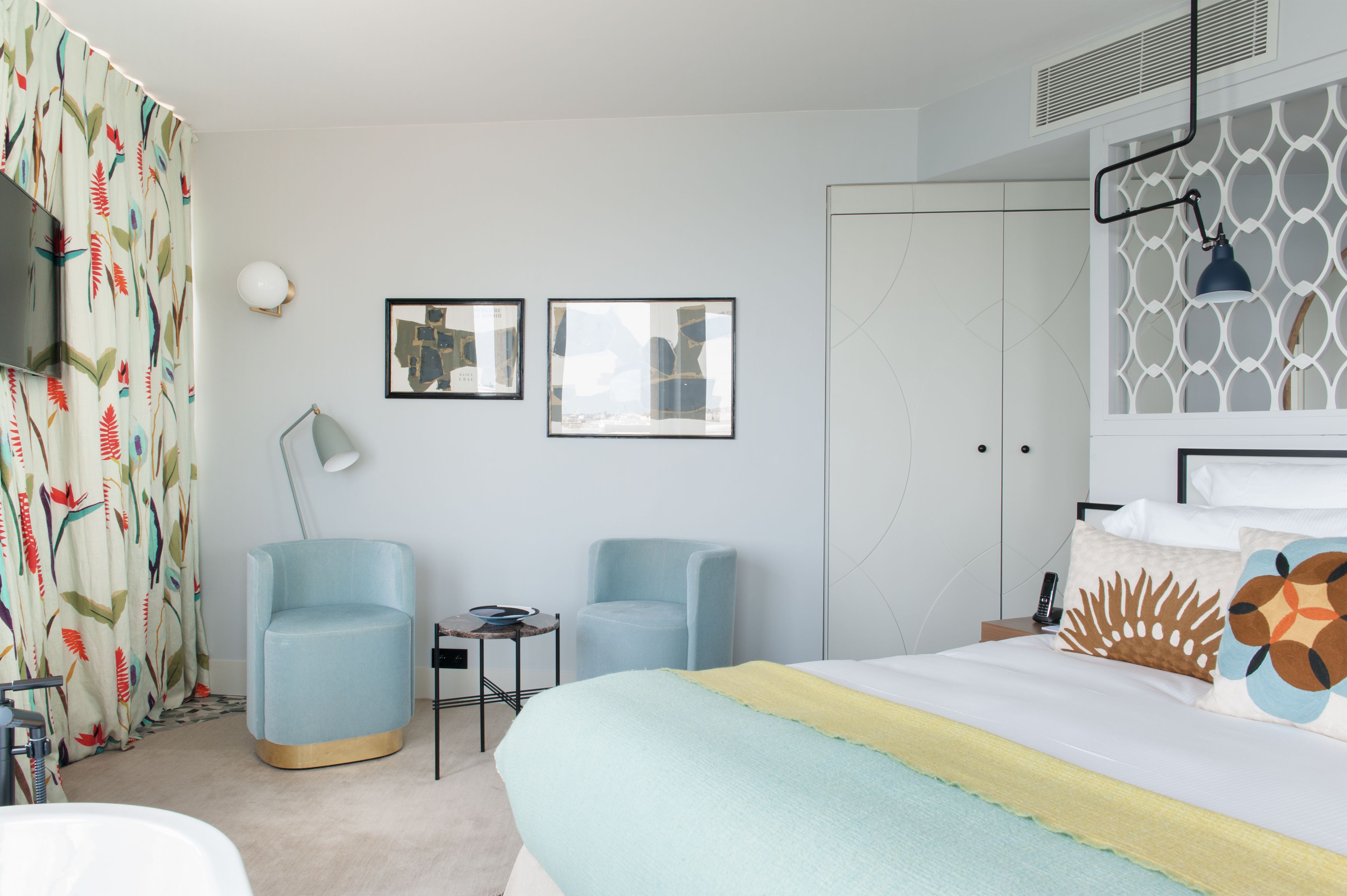 Une chambre d’hôtel moderne, équipée d’un lit, de deux fauteuils, d’une petite table, d’un téléviseur et d’œuvres d’art accrochées au mur.