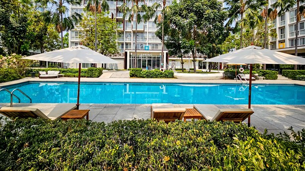 A swimming pool with lounge chairs and umbrellas, surrounded by greenery and a multi-story building with balconies. - The Idle Hotel and Residence (Khlong Luang)