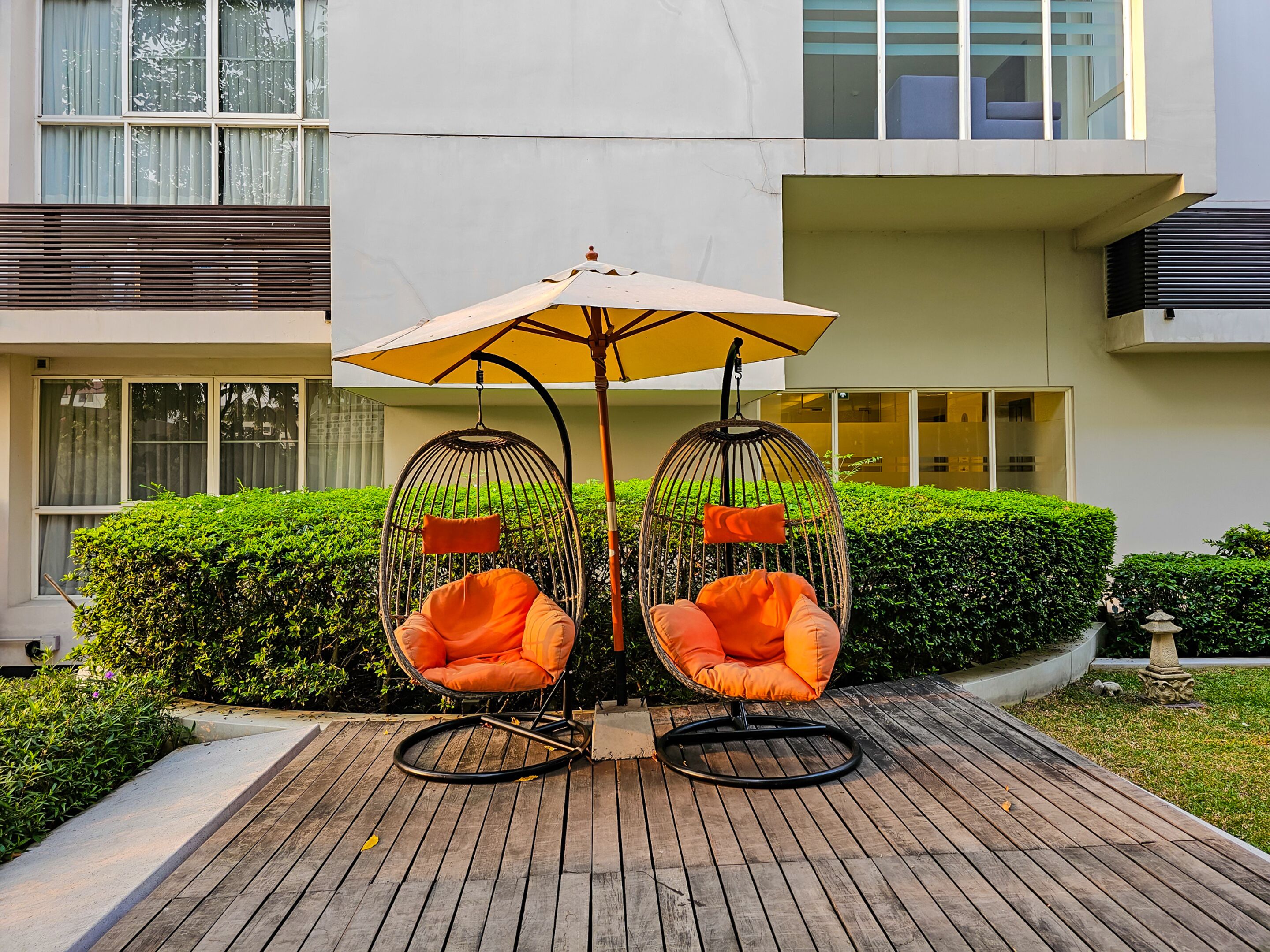 A wooden deck with two wicker chairs under a large umbrella, surrounded by well-maintained hedges and a building in the background.