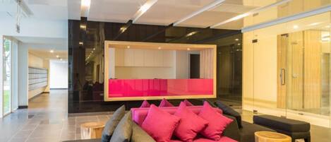 A modern lobby with a grey sofa and pink cushions, a wooden coffee table, and a glass partition.