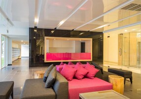 A modern lobby with a grey sofa and pink cushions, a wooden coffee table, and a glass partition.