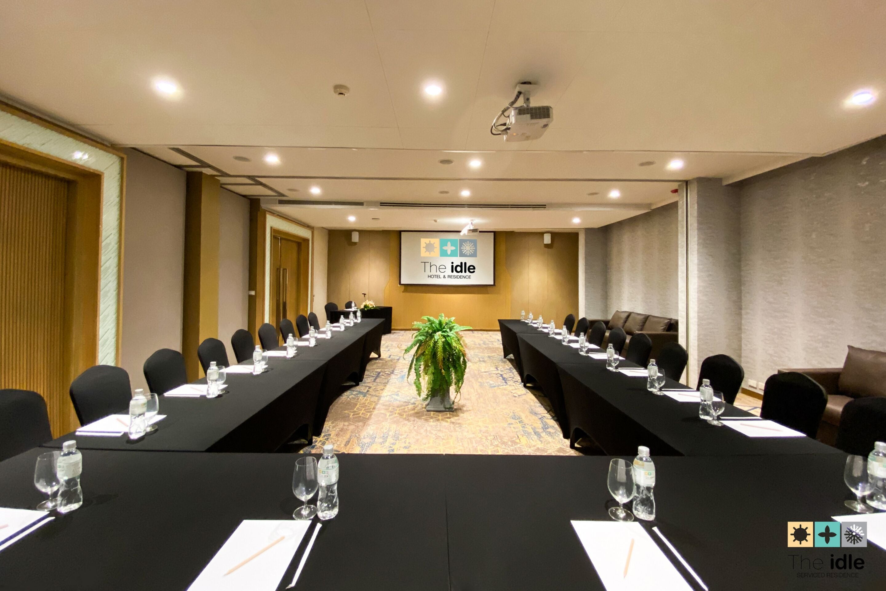 A conference room with long tables, chairs, a projector screen, and a potted plant.