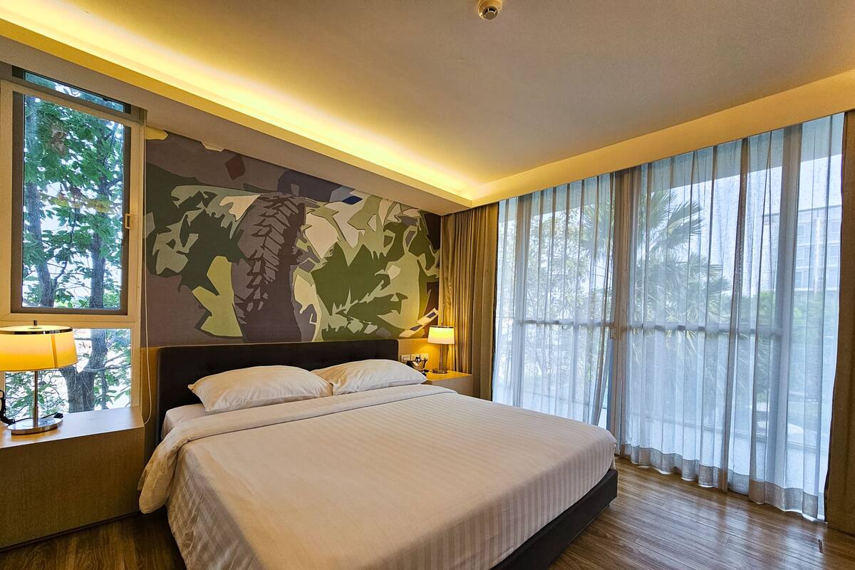 Una habitació d'hotel amb un llit gran, una nitòria amb una làmpada, una paret amb dibuixos de camuflatge i vista a arbres a través de cortines primes.