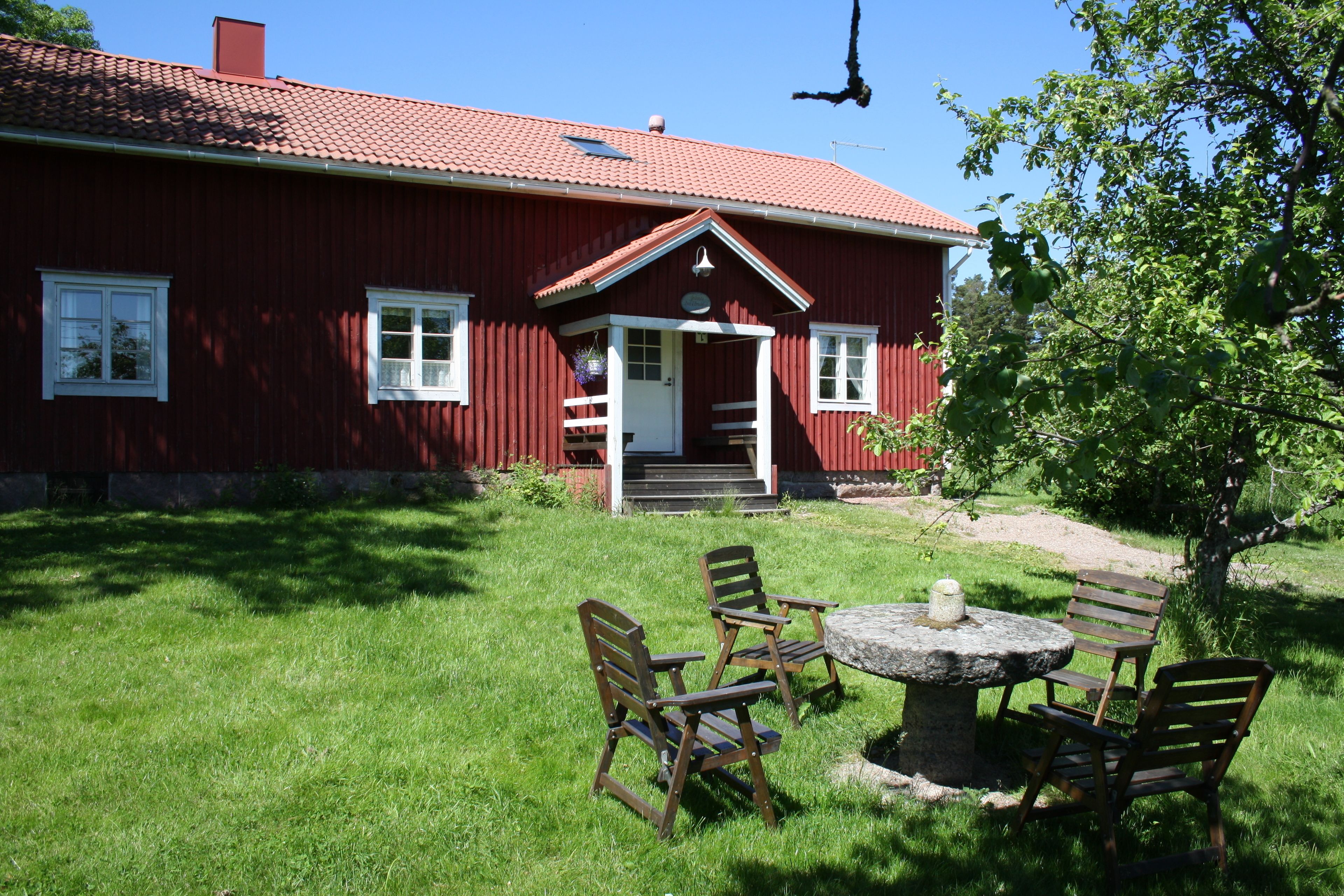 A red wooden house with a tiled roof, a front porch with steps, and a stone table with chairs in the yard.