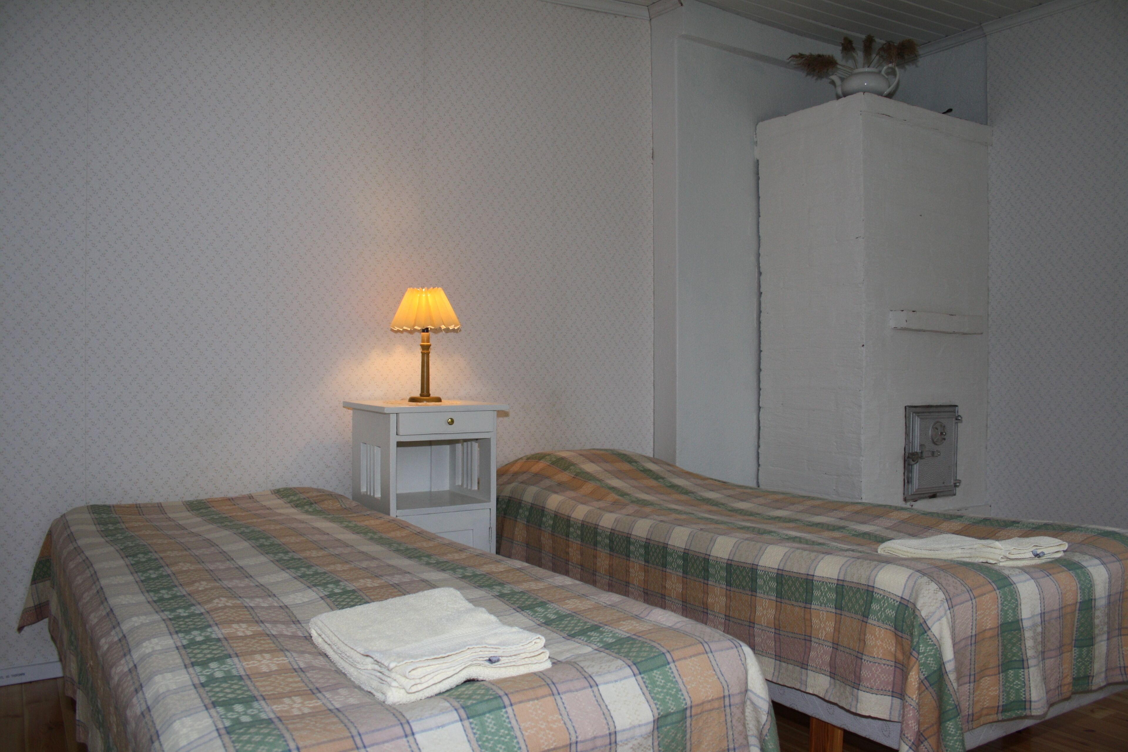 A room with two beds, each with a plaid bedspread and a folded towel on the bed. There is a nightstand with a lamp between the beds.
