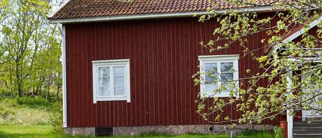 Une maison en bois rouge avec un toit en tuiles, un petit jardin avec une table ronde en pierre et deux chaises en bois, et une clôture en bois au premier plan.