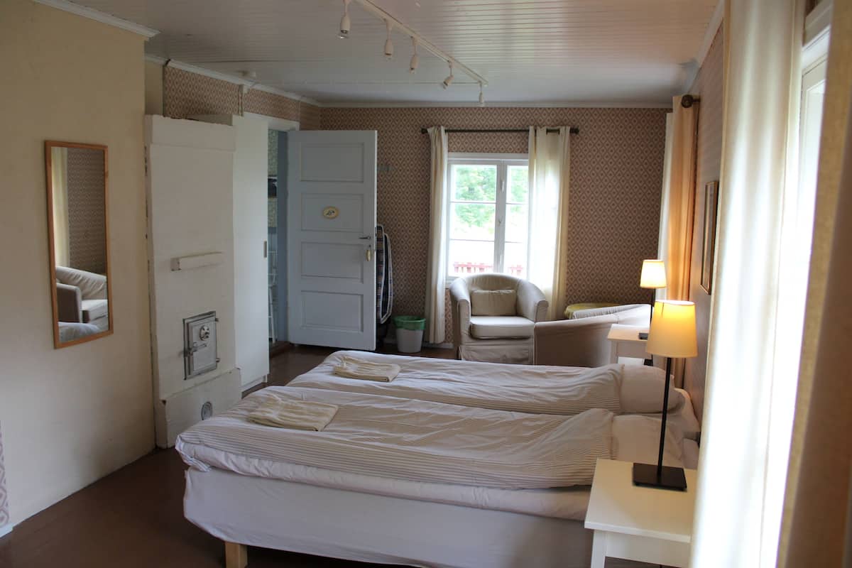 Ein Hotelzimmer mit zwei Betten, einem Sessel, einem Schreibtisch, einer Lampe und einem Fenster mit Vorhängen.