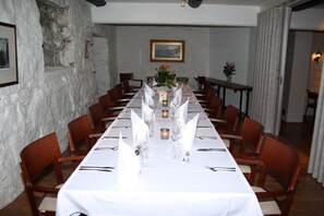 Una larga mesa de comedor con manteles blancos, velas y copas, rodeada de sillas de madera en una habitación con paredes de piedra y un cuadro colgado.