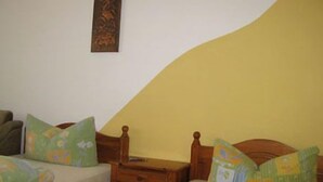 Un dormitorio con dos camas individuales, cada una con cabecero de madera y ropa de cama estampada con motivos florales verdes. Hay una mesita de noche de madera entre las camas, y un pequeño aparador a la izquierda.