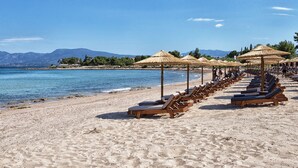 Une plage bordée de rangées de chaises longues et d’ombrelles en chaume, un ciel bleu azur et des montagnes en arrière-plan.