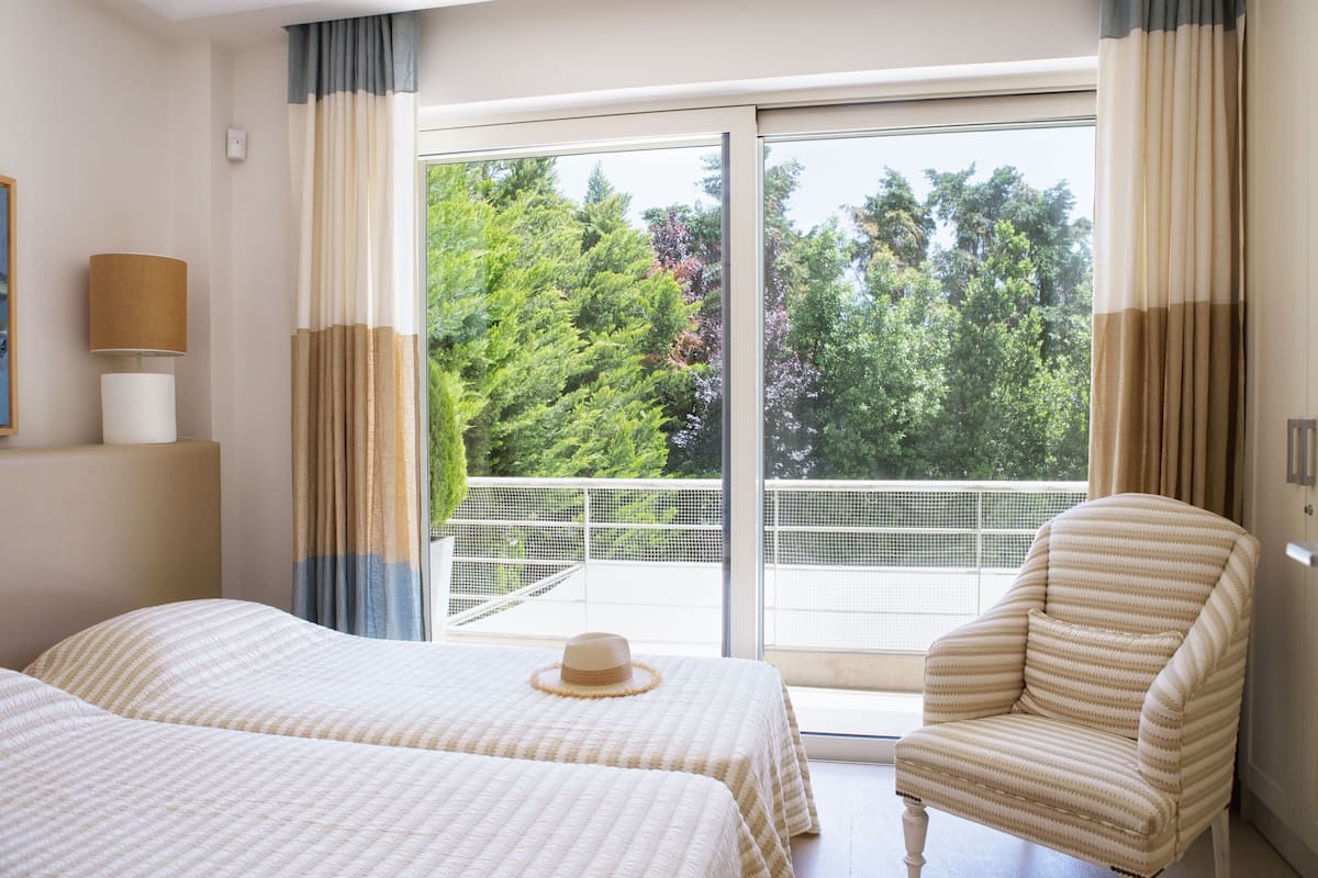 A hotel room with two beds, a chair, a hat on one bed, and a balcony with a view of trees.