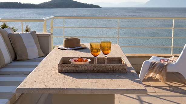Une terrasse avec une table dressée pour deux personnes, agrémentée d’un plateau en osier contenant des oranges et des verres, donnant sur une étendue d’eau avec une terre ferme visible au loin.