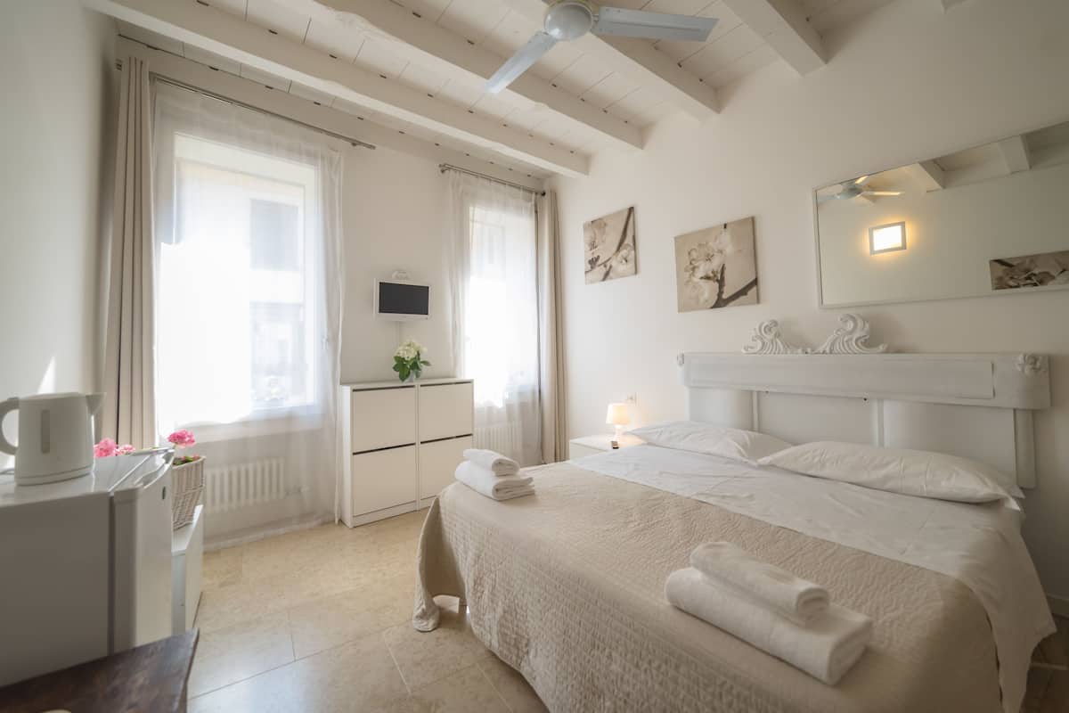 Ein Hotelzimmer mit einem Bett, einem Schreibtisch, einem Stuhl, einem Heizkörper, einem Fenster mit Vorhängen, einem an der Wand befestigten Fernseher und Kunstwerken.