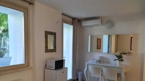 A hotel room with a bed, a desk with two chairs, a small refrigerator, a microwave, a radiator, an air conditioning unit, and a window with a view of greenery.