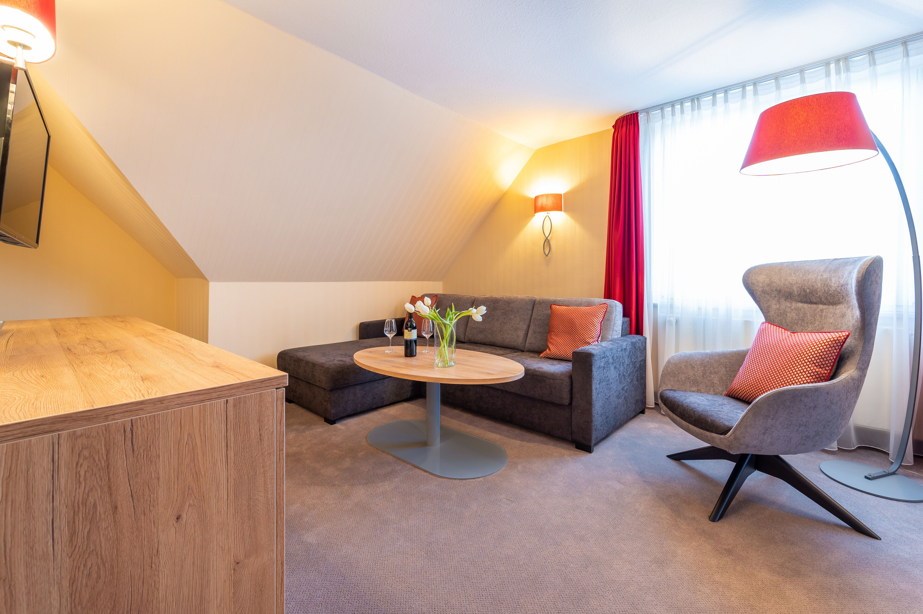 A modern hotel room with a wooden desk, a round coffee table with a vase, a grey sofa with red cushions, a grey armchair, and a red floor lamp.