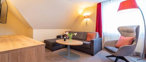 Una habitación de hotel moderna con un escritorio de madera, una mesa redonda de centro con un florero, un sofá gris con cojines rojos, un sillón gris y una lámpara de pie roja.