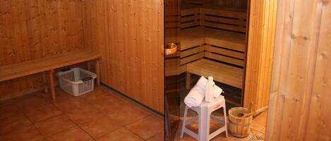 A sauna room with wooden walls and benches. There is a bench with a basket underneath and a stool with towels on it.